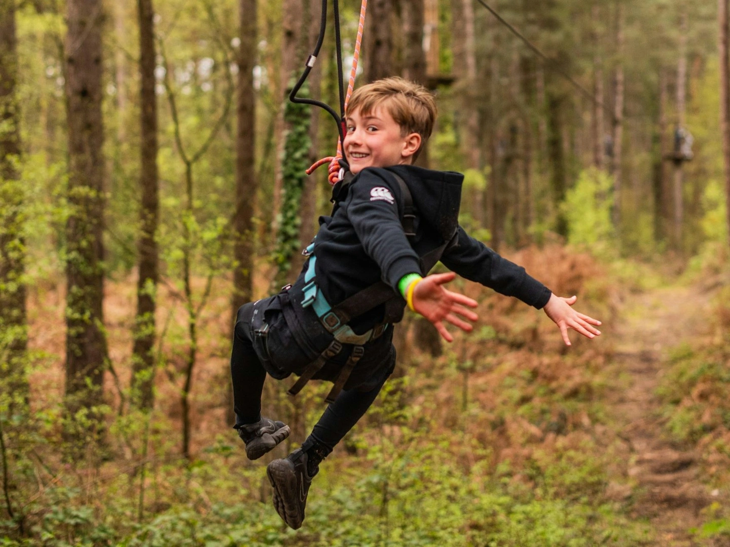 A boy with his arms stretched out and smiling as he rides a zip wire through the forest