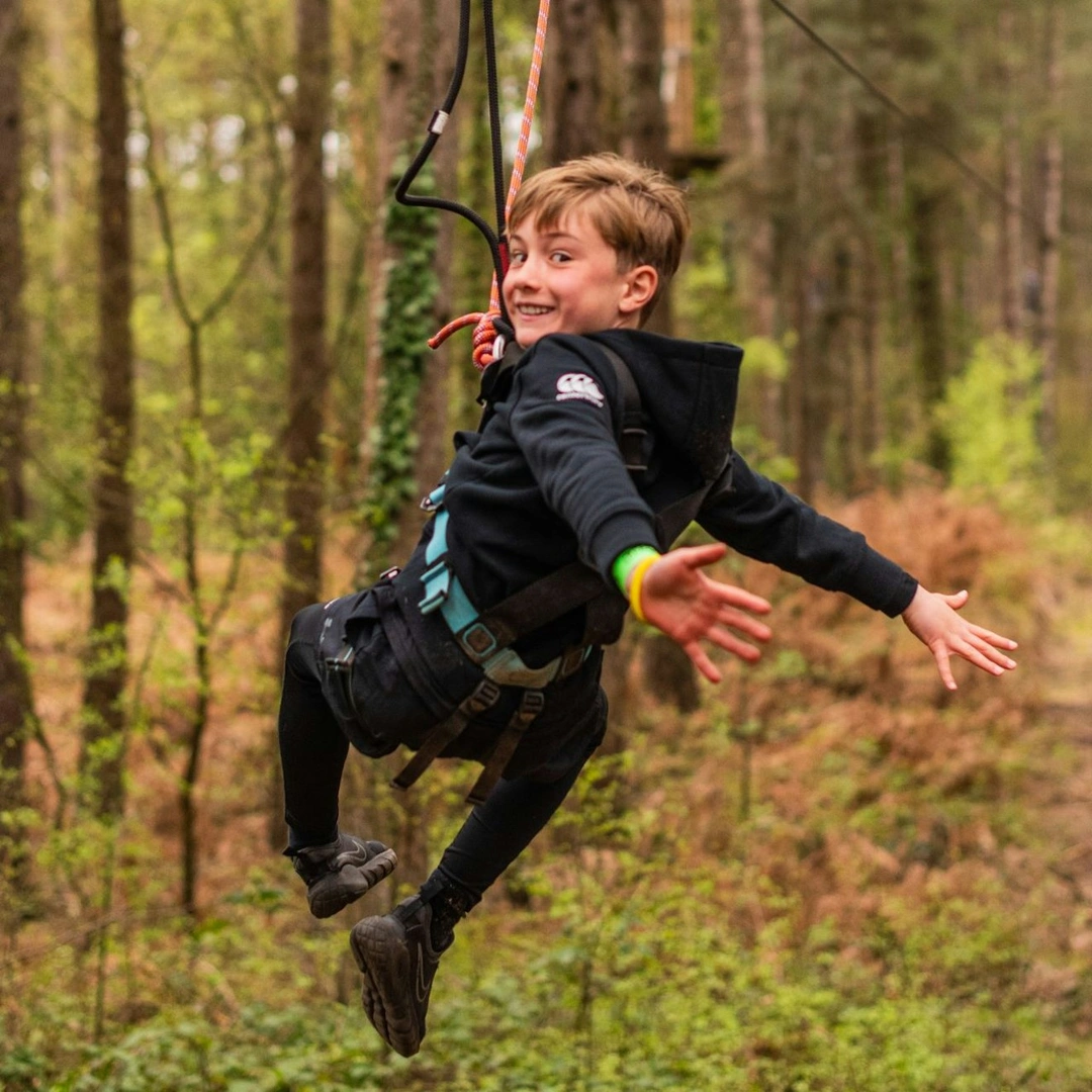 A boy with his arms stretched out and smiling as he rides a zip wire through the forest