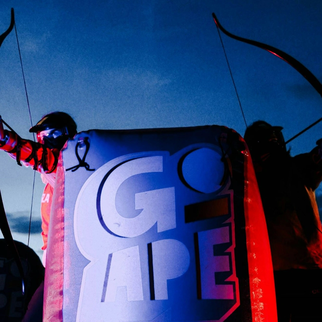 Two people draw Archery Tag bows either side of an inflatable Go Ape branded barricade