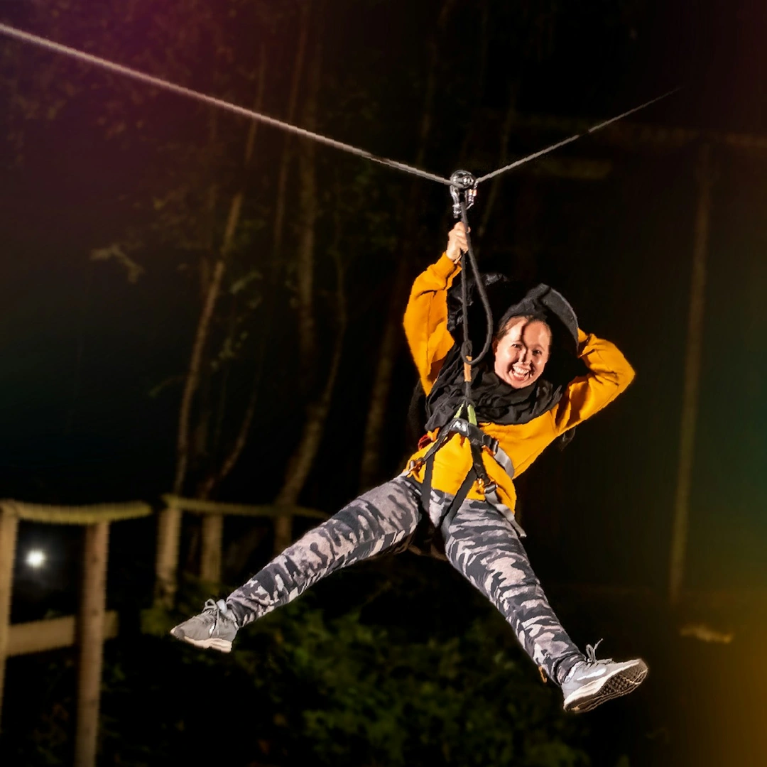 A woman rides a zip wire after dark surrounded by colourful lights